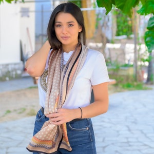 May include: A woman wearing a white t-shirt with a brown and black patterned scarf. The scarf has a gold logo that says "VERSACE". She is wearing blue jeans and has her hair pulled back.