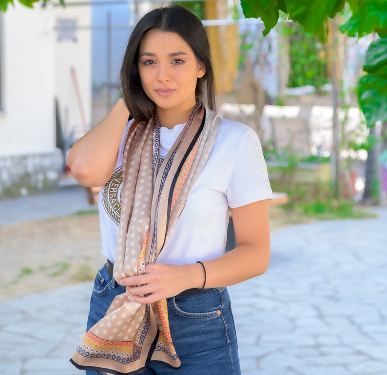 May include: A woman wearing a white t-shirt with a brown and black patterned scarf. The scarf has a gold logo that says "VERSACE". She is wearing blue jeans and has her hair pulled back.