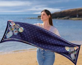 May include: A woman wearing blue jeans and a white top holds a blue silk scarf with a colorful pattern of circles. The scarf has a border of small, colorful squares.