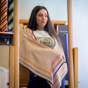 May include: A woman wearing a white t-shirt with a logo that says "Versace" and a beige scarf with a polka dot pattern and a colorful border. The scarf is wrapped around her neck and shoulders.
