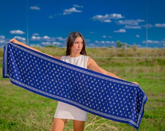 May include: A woman holds a blue scarf with a white pattern of the Greek alphabet. The scarf is decorated with a white border. The woman is wearing a white sleeveless dress and is standing in a field of green grass.