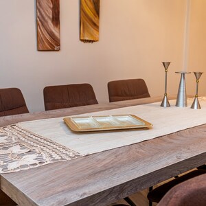 May include: A wooden dining table with a white tablecloth and a crocheted runner. There are three silver candle holders with gold accents on the table. Four brown chairs surround the table.