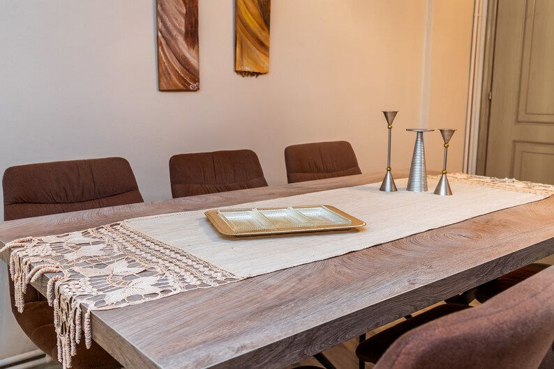 May include: A wooden dining table with a white tablecloth and a crocheted runner. There are three silver candle holders with gold accents on the table. Four brown chairs surround the table.