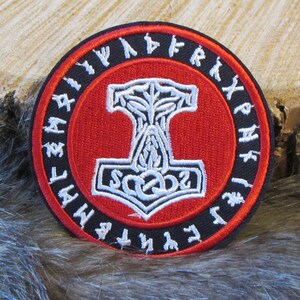 May include: A red and black embroidered patch with a white Mjolnir hammer surrounded by a circle of white runes. The runes are in a circle around the hammer and are in a black font.