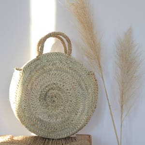 May include: A round, woven straw bag with two handles. The bag is light beige and has a circular shape. Two tall, dried beige ornamental grasses are next to the bag. The background is a white wall.