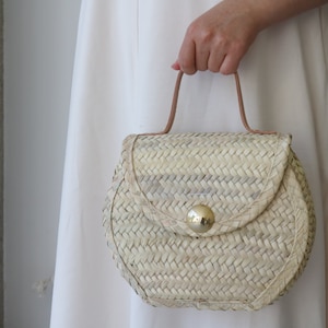 May include: A woven straw handbag with a rounded shape and a brown leather handle. The bag has a gold metal clasp.
