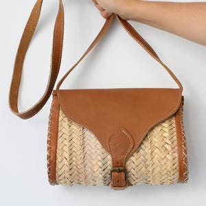 May include: A brown leather and woven straw crossbody bag with a flap closure and a buckle. The bag has a long adjustable strap.