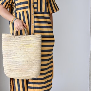 Pode incluir: Uma sacola de palha tecida com formato retangular e duas alças. A sacola é segurada por uma pessoa que veste um vestido camisa listrado amarelo e azul marinho. O vestido tem uma frente abotoada e mangas curtas.