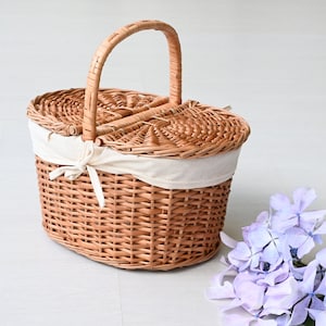 Pode incluir: Uma cesta de piquenique de vime trançado com um forro de tecido branco e uma alça marrom. A cesta é oval e tem uma tampa com dobradiças.