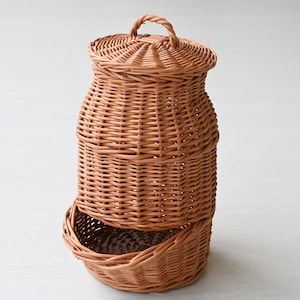 May include: A woven wicker basket with a lid and a lower, open basket section. The basket is a warm brown color and has a handle on the lid. The basket is designed for storage and organization.
