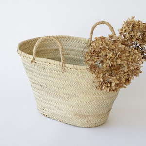 May include: A woven straw tote bag with two handles. The bag is oval-shaped and has a natural, light tan color. Dried brown flowers are placed inside the bag, adding a decorative touch. The bag is suitable for carrying items.