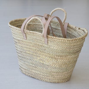 May include: Woven straw tote bag with light brown leather handles. The bag has a natural, light tan color and a classic oval shape. The handles are attached with small, light pink leather accents. The bag is empty and sits on a light gray surface.