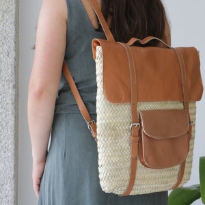 May include: A woven straw backpack with brown leather accents. The backpack has a large flap with a buckle closure and a smaller pocket on the front.
