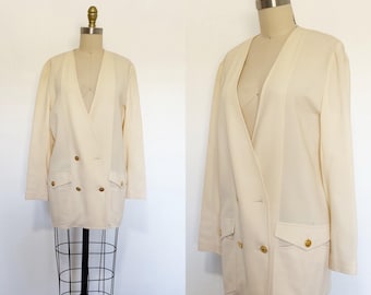 May include: A cream-coloured double-breasted blazer featuring a deep V-neckline, long sleeves, and two front flap pockets. The garment is accented with gold-toned buttons on the front closure and on the pocket flaps, displayed on a dress form.