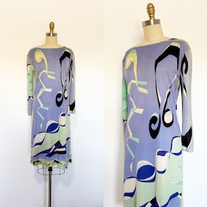 May include: A long-sleeve dress featuring a light purple base with a graphic pattern of black, white, blue, and pale green abstract shapes. The garment has a boat neckline and a straight silhouette, displayed on a mannequin for full visibility.