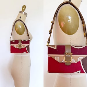 May include: A rectangular shoulder bag featuring a geometric pattern in shades of deep red, brown, and beige. The bag has a tan leather strap with gold-tone hardware studs and a central tan leather flap closure secured by a gold-tone metal buckle.
