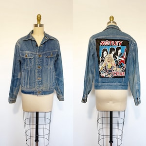 May include: A blue denim jacket featuring a pointed collar, long sleeves, and a button-up front. The back displays a large rectangular patch with a graphic of the band Mötley Crüe, including band members and the text Mötley Crüe in red and white.