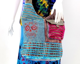 Pode incluir: Bolsa de ombro colorida em patchwork com um design de caveira do Dia dos Mortos. A bolsa apresenta tecido jeans, castanho e rosa com vários bolsos e uma alça longa. A bolsa é exibida em um manequim vestindo uma camisa tie-dye e uma saia azul.