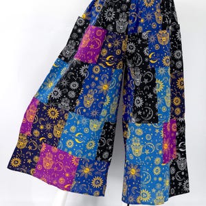 Puede incluir: Pantalones anchos de patchwork con un dise&ntilde;o celestial. Los pantalones presentan una mezcla de paneles azules, negros, morados y amarillos, cada uno adornado con soles, lunas, estrellas y motivos de manos de Hamsa. La cintura es de color morado liso.
