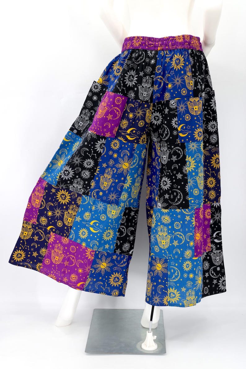 Puede incluir: Pantalones anchos de patchwork con un dise&ntilde;o celestial. Los pantalones presentan una mezcla de paneles azules, negros, morados y amarillos, cada uno adornado con soles, lunas, estrellas y motivos de manos de Hamsa. La cintura es de color morado liso.