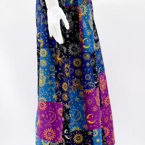 Puede incluir: Una falda larga de patchwork con s&iacute;mbolos celestiales y espirituales. La falda tiene una cintura morada y est&aacute; compuesta por varios paneles rectangulares en azul, negro y morado, adornados con soles, lunas, estrellas y motivos de manos. La falda se exhibe en un maniqu&iacute;.