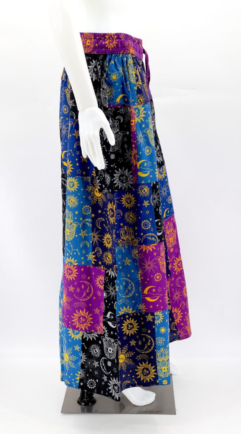 Puede incluir: Una falda larga de patchwork con s&iacute;mbolos celestiales y espirituales. La falda tiene una cintura morada y est&aacute; compuesta por varios paneles rectangulares en azul, negro y morado, adornados con soles, lunas, estrellas y motivos de manos. La falda se exhibe en un maniqu&iacute;.