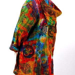 K&ouml;nnte beinhalten: Eine bunte Kapuzenjacke mit Batik-Mustern und schwarzen, gedruckten Symbolen, darunter Sonnen, Monde und Blumen.