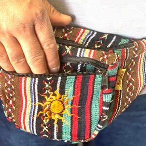 K&ouml;nnte beinhalten: Eine schwarz-braun-rot-blau gestreifte Bauchtasche mit einem Rei&szlig;verschlussfach und einem gestickten Sonnendesign. Die Bauchtasche hat ein braunes Leder-Etikett mit dem aufgedruckten Wort "Himalaya".