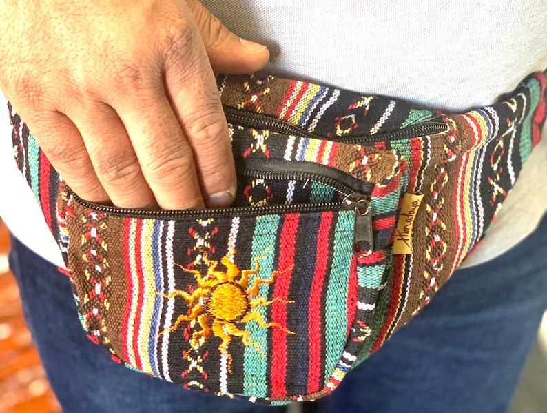 K&ouml;nnte beinhalten: Eine schwarz-braun-rot-blau gestreifte Bauchtasche mit einem Rei&szlig;verschlussfach und einem gestickten Sonnendesign. Die Bauchtasche hat ein braunes Leder-Etikett mit dem aufgedruckten Wort "Himalaya".
