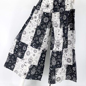 Puede incluir: Pantalones palazzo de patchwork en blanco y negro con dise&ntilde;os celestiales y en forma de mano. Los pantalones de pernera ancha presentan una mezcla de cuadrados con soles, lunas, estrellas y el s&iacute;mbolo de la mano de Hamsa. Los pantalones se muestran en un maniqu&iacute;.