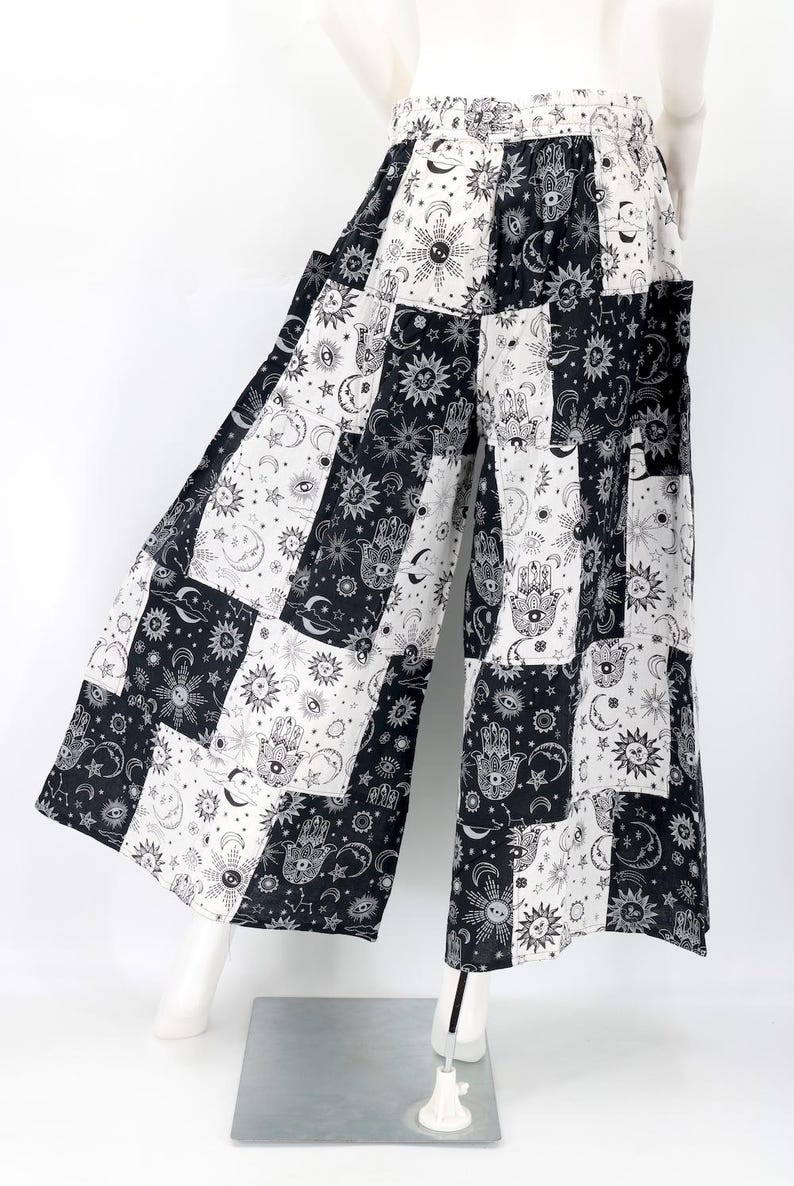 Puede incluir: Pantalones palazzo de patchwork en blanco y negro con dise&ntilde;os celestiales y en forma de mano. Los pantalones de pernera ancha presentan una mezcla de cuadrados con soles, lunas, estrellas y el s&iacute;mbolo de la mano de Hamsa. Los pantalones se muestran en un maniqu&iacute;.
