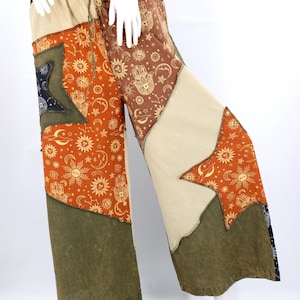 Op de afbeelding: Patchwork broek met wijde pijpen, elastische tailleband en trekkoord. De broek heeft verschillende stoffen panelen in tinten oranje, bruin en olijfgroen, versierd met hemelse en handmotieven. Gepresenteerd op een mannequin.