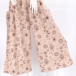 Op de afbeelding: Zandkleurige palazzo broek met een all-over print van hemelse symbolen, waaronder zonnen, manen, sterren en Hamsa-handen. De broek met wijde pijpen heeft zakken en een elastische tailleband. De stof is licht.