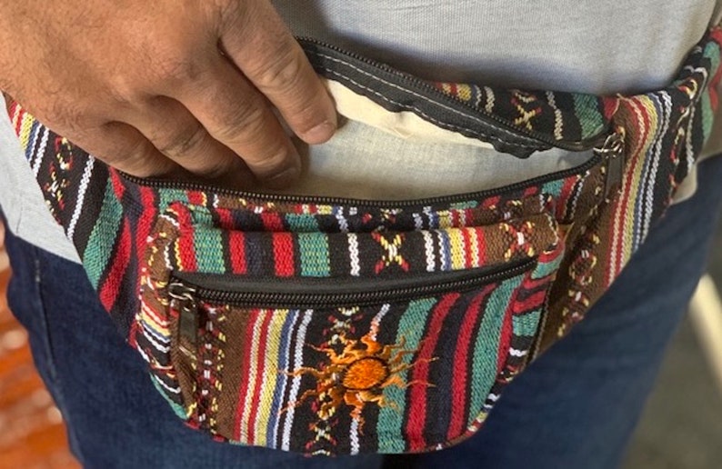 K&ouml;nnte beinhalten: Eine mehrfarbige, gestreifte Bauchtasche mit einem Rei&szlig;verschlussfach und einem Sonnendesign. Die Bauchtasche ist aus gewebtem Stoff und hat einen schwarzen Rei&szlig;verschluss.