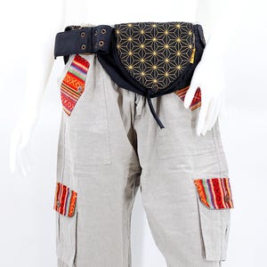 May include: A black canvas belt with a geometric pattern fabric pouch. The pouch has a drawstring closure and is attached to the belt with two metal grommets. The belt is worn over a pair of beige cargo pants with colorful patterned fabric accents on the pockets.