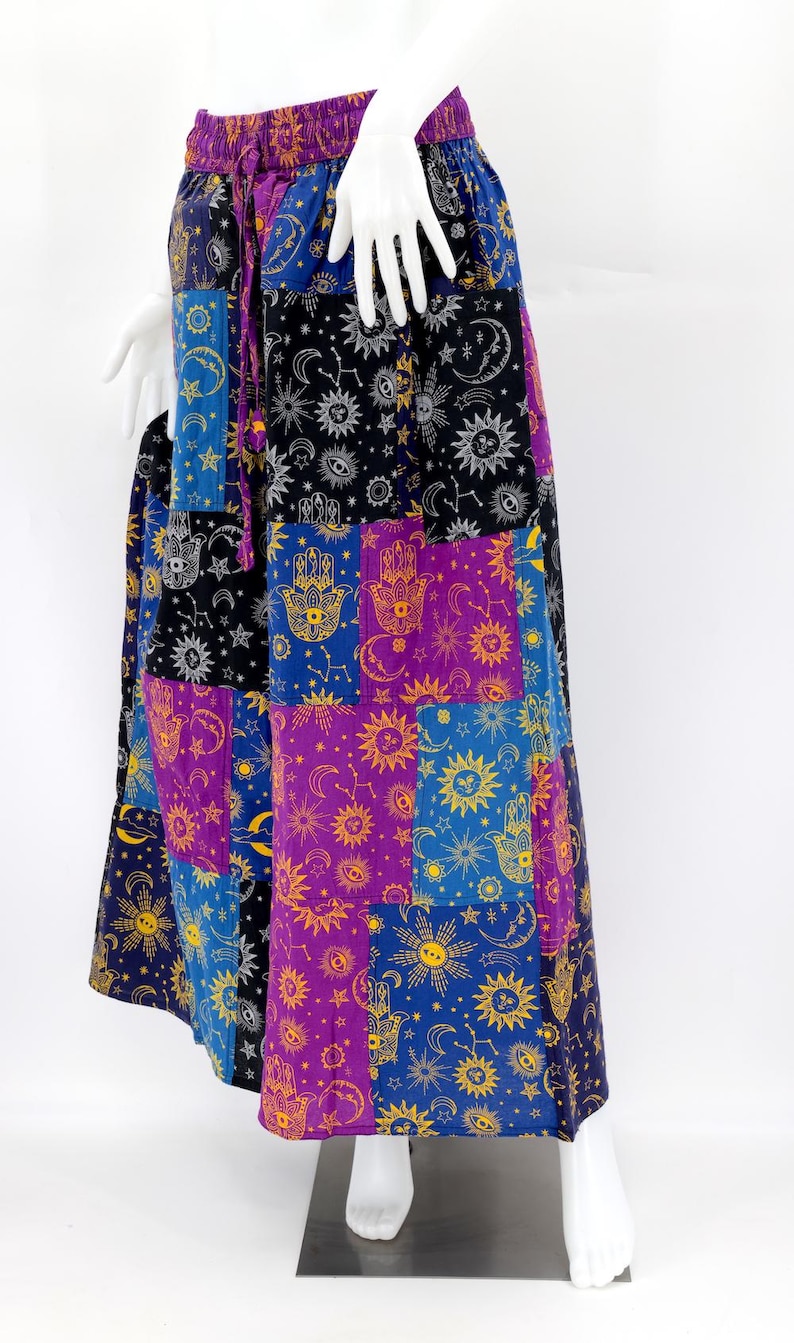 Puede incluir: Falda larga de patchwork con dise&ntilde;o celestial y mano de F&aacute;tima. La falda tiene una cintura morada con cord&oacute;n y est&aacute; compuesta por cuadrados en tonos azules, morados, negros y dorados. La falda se exhibe en un maniqu&iacute;.