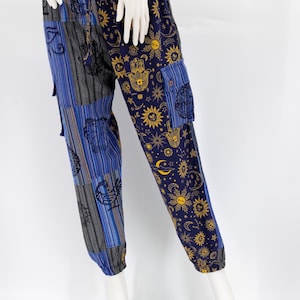May include: Patchwork pants with a navy blue background and gold sun, moon, and hand patterns. The pants have blue and gray striped panels and blue pockets. The pants are displayed on a white mannequin.