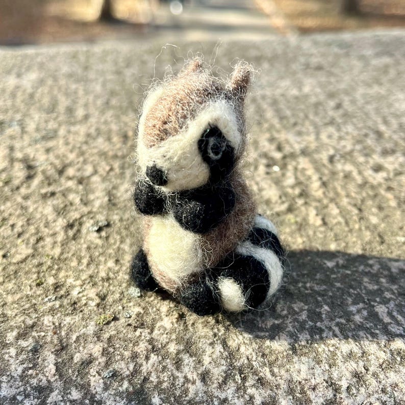 Peut inclure: Une petite figurine de raton laveur en feutre, faite &agrave; la main, avec un motif brun, blanc et noir. Le raton laveur est assis droit, les pattes repli&eacute;es devant lui.