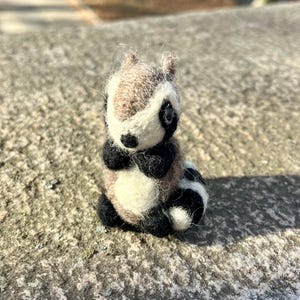Peut inclure: Une petite figurine de raton laveur en feutre, faite &agrave; la main, avec une fourrure brune, blanche et noire. Le raton laveur est assis droit, les pattes repli&eacute;es devant lui.