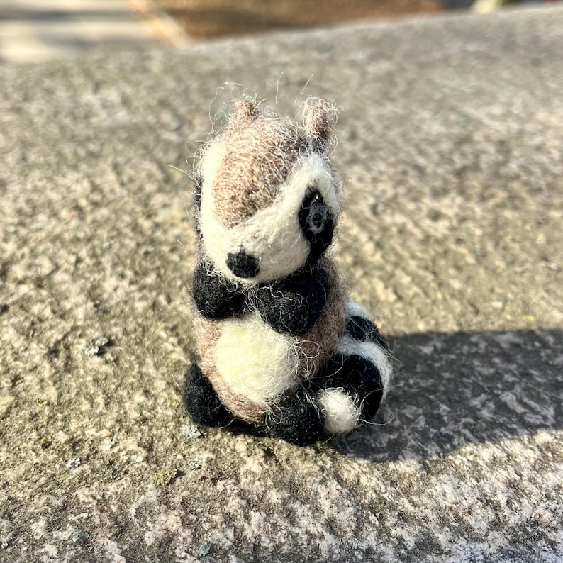 Peut inclure: Une petite figurine de raton laveur en feutre, faite &agrave; la main, avec une fourrure brune, blanche et noire. Le raton laveur est assis droit, les pattes repli&eacute;es devant lui.