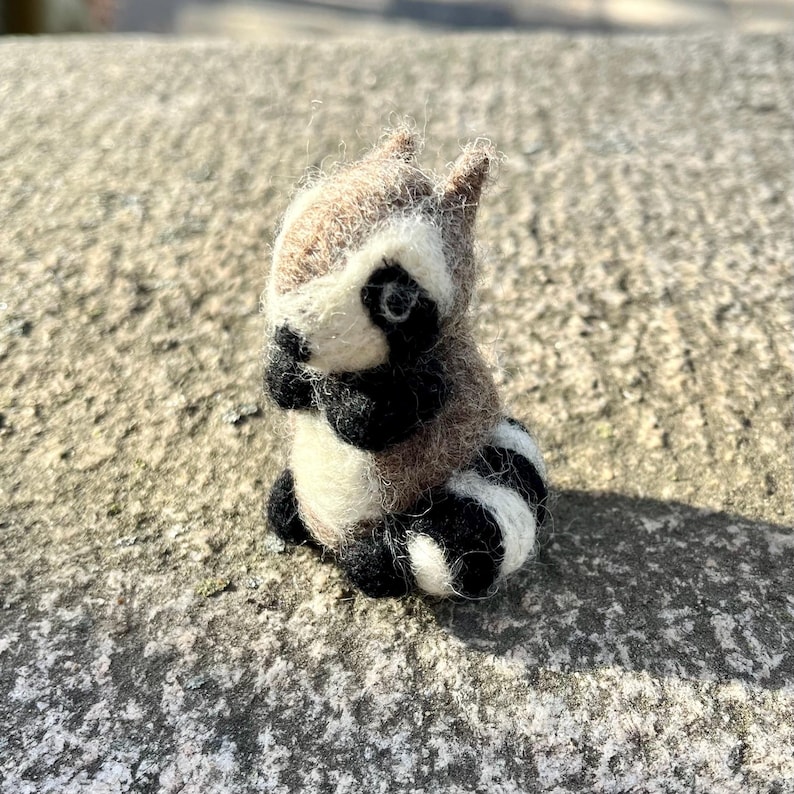 Peut inclure: Une petite figurine de raton laveur en feutre, faite &agrave; la main, avec des marques noires et blanches. La figurine est en laine et a un aspect r&eacute;aliste.