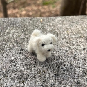 May include: A small, white, felted dog figurine with black eyes. The figurine is sitting on a gray, textured surface.