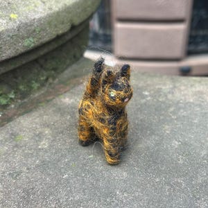 Pode incluir: Uma pequena figura de gato feita à mão de feltro, com pelo preto e amarelo. O gato está em pé sobre suas quatro patas com as patas dianteiras levantadas.