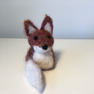 May include: A handcrafted felt fox figurine. The fox has a brown and white body with a fluffy tail. The fox has black eyes and a black nose. The fox is sitting on a white surface.