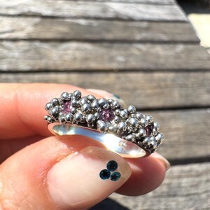 May include: A silver ring featuring a textured, bubbly surface composed of numerous small, rounded metallic spheres. Embedded within this granular, organic-looking metal structure are three small, round, faceted pink gemstones spaced evenly apart.