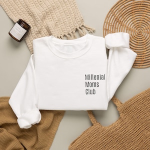 May include: A white crewneck sweatshirt featuring the black text Millenial Moms Club printed on the left chest area. The garment has long sleeves with ribbed cuffs and a ribbed neckline, presented in a clean, simple design for everyday wear.