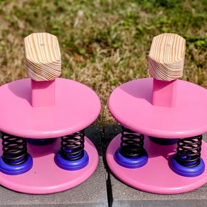 May include: Two pink wooden spring stools with a circular base and top. Each stool has a wooden block on top, black springs, and blue accents. The stools are designed for children's play and are placed on a grey surface.