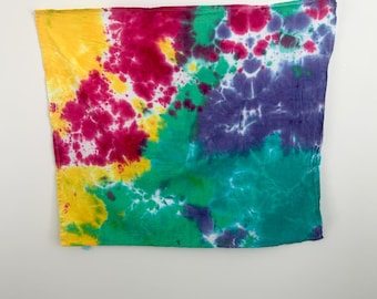May include: A tie-dye textile with a vibrant color scheme. The fabric features a blend of yellow, fuchsia, green, teal, and purple hues. The dye pattern creates a unique, abstract design. The textile is rectangular and appears to be made of a soft material.