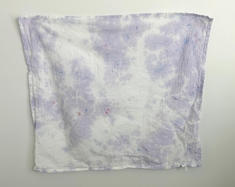May include: A square textile cloth featuring a tie-dye pattern in shades of light purple and white. The fabric displays a soft, mottled appearance with subtle, scattered flecks of blue and red pigment throughout the surface of the material.