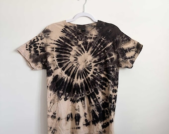 May include: A short-sleeve t-shirt featuring a black and beige tie-dye spiral pattern. The design radiates from the center of the chest, creating a bold contrast between the dark black ink and the light beige fabric across the entire garment.
